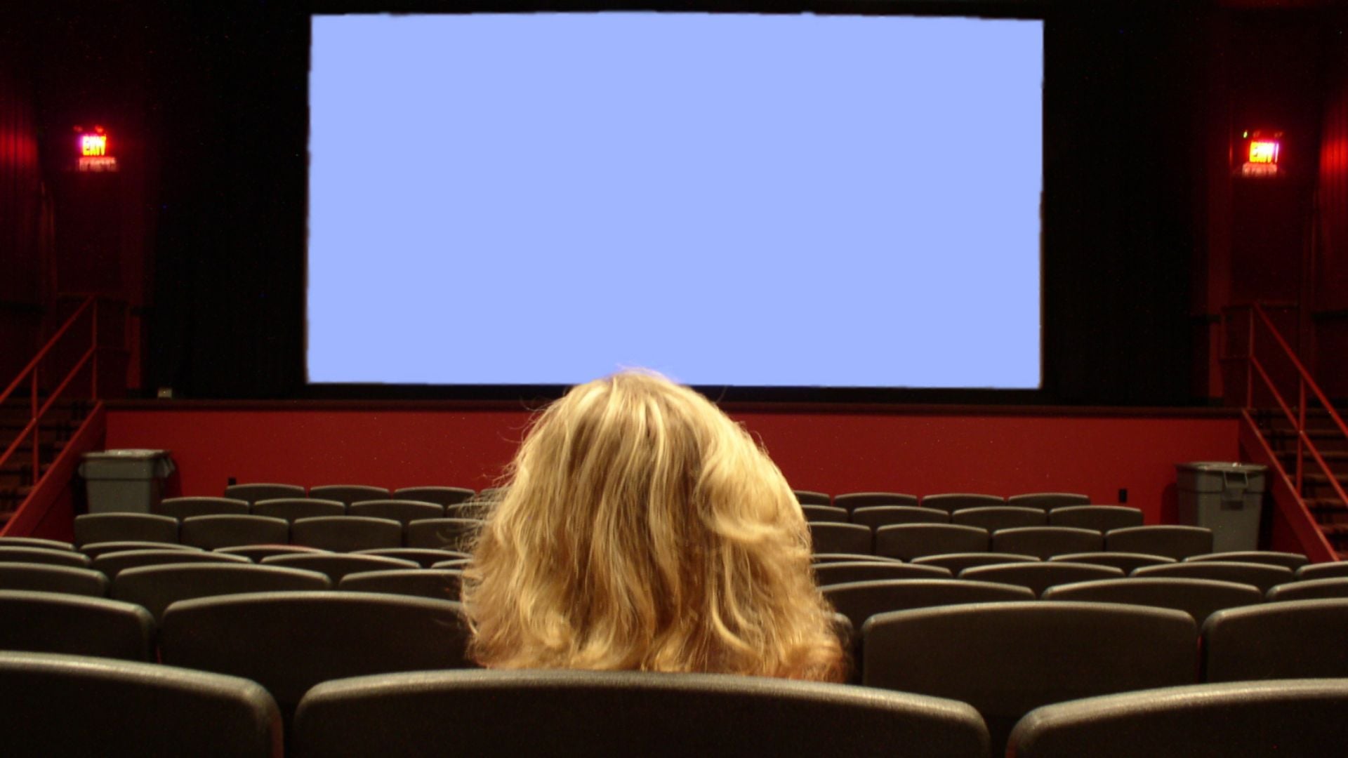 A person with blonde hair sits alone in a mostly empty movie theater, facing a large blank screen with rows of black seats around them.
