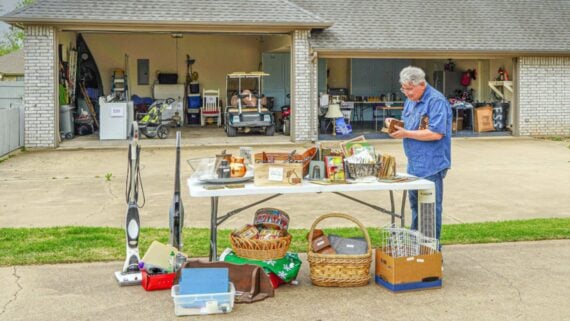 A man browses items on a table at a garage sale in front of a suburban house with an open garage, showing various household goods for sale, including baskets, appliances, and knickknacks.