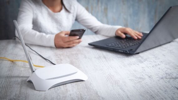 A person sits at a table using a laptop with one hand and holding a smartphone in the other. A white Wi-Fi router with antennas is in the foreground on the wooden surface.