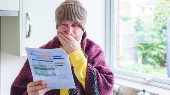 A person wearing a brown beanie and wrapped in a blanket stands in a kitchen by a window, looking concerned while holding and reading a bill or statement.