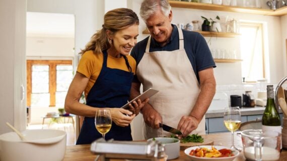 A woman and an older man, both wearing aprons, cook together in a bright kitchen. The woman smiles while holding a tablet, and the man chops herbs. Wine glasses and food are on the counter beside them.