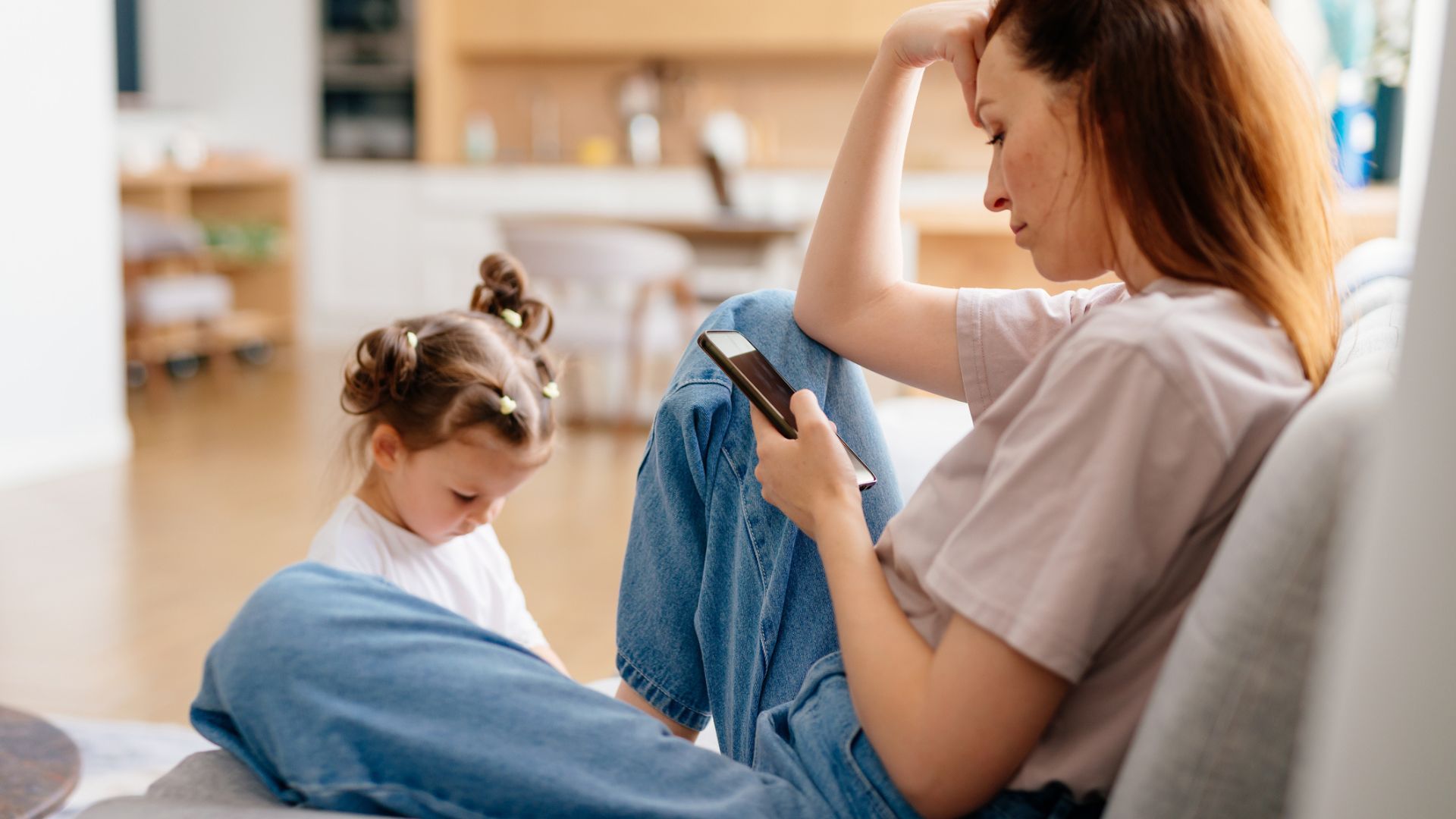 A woman sits on a couch looking stressed while holding her phone, with her elbow on her knee. Next to her, a young girl with two buns in her hair sits on the floor, playing and looking down.