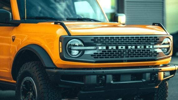 Close-up of the front of a bright yellow Ford Bronco, showing the grille with "BRONCO" lettering, round headlights, rugged tires, and part of the hood and bumper.