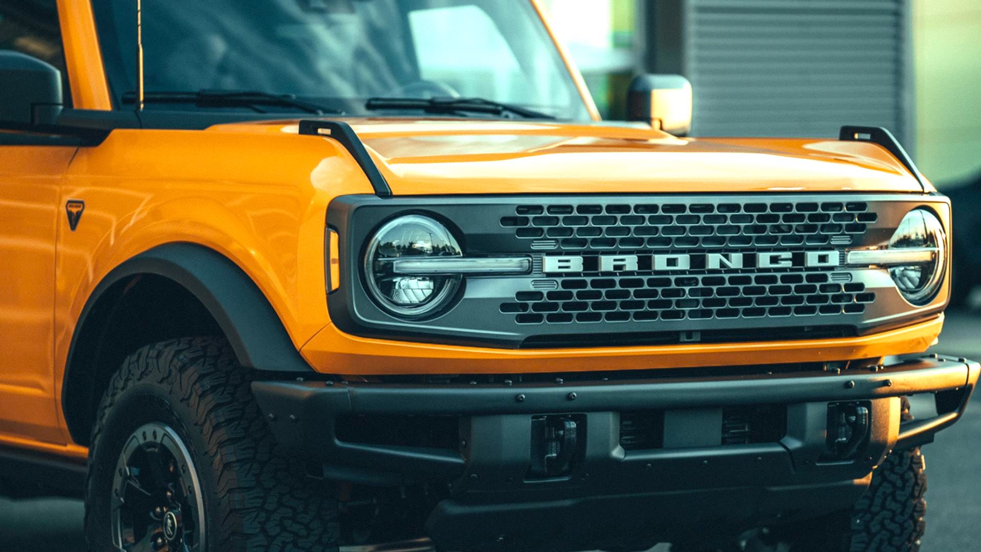 Close-up of the front of a bright yellow Ford Bronco, showing the grille with "BRONCO" lettering, round headlights, rugged tires, and part of the hood and bumper.