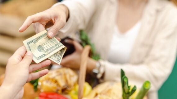 A person hands over U.S. dollar bills to another person, with fresh produce and groceries visible in the foreground. The background is softly blurred.