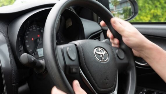 Close-up of a person’s hands holding a Toyota steering wheel inside a car, with part of the dashboard, speedometer, and side mirror visible in the background.