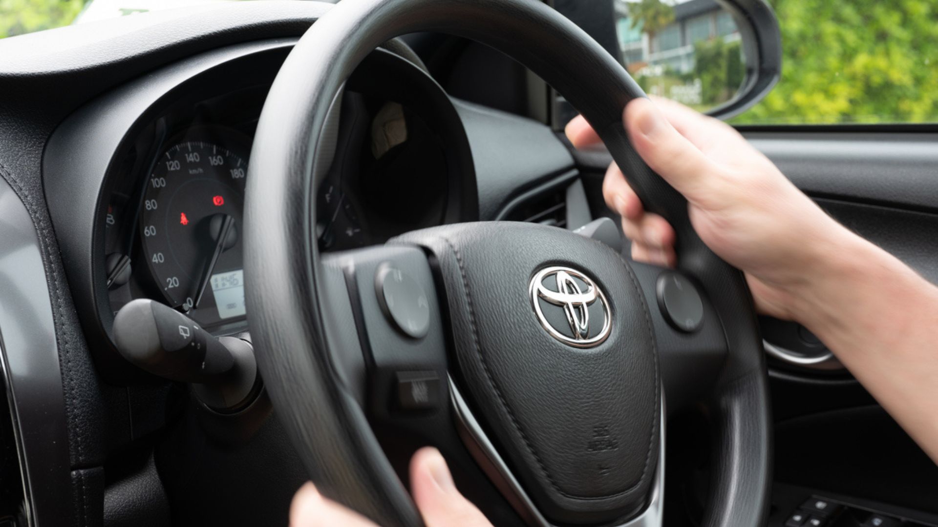 Close-up of a person’s hands holding a Toyota steering wheel inside a car, with part of the dashboard, speedometer, and side mirror visible in the background.