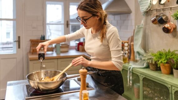A woman wearing glasses and a white top pours a dark liquid from a bottle into a pan while cooking in a bright, modern kitchen with plants and utensils on the counter.