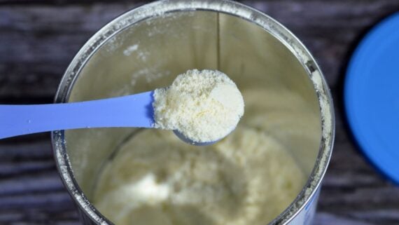 A close-up of a blue plastic scoop holding powdered infant formula above an open metal container filled with the same powder, with a blue lid placed beside the container.