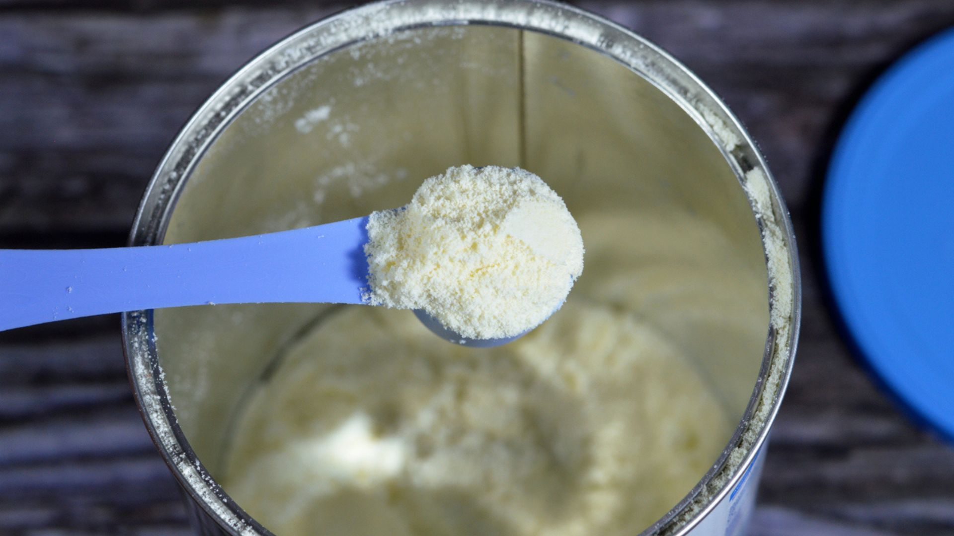 A close-up of a blue plastic scoop holding powdered infant formula above an open metal container filled with the same powder, with a blue lid placed beside the container.