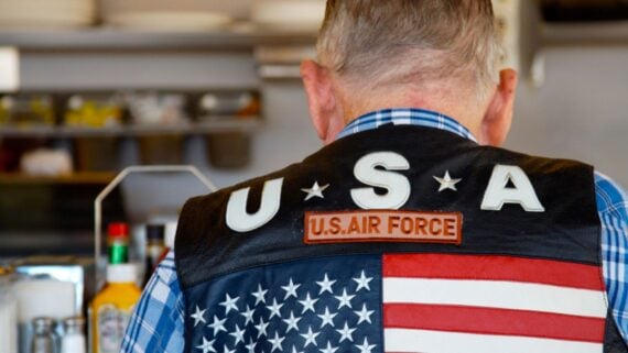 A man wearing a blue plaid shirt and a leather vest with "U.S.A," "U.S. Air Force," and an American flag embroidered on the back is sitting at a counter, seen from behind.