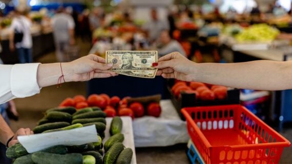 Two people exchange U.S. dollar bills over a vegetable stand at a market, with cucumbers and tomatoes visible in the foreground and blurred people and produce in the background.