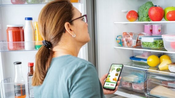 A woman with glasses looks into an open refrigerator while holding a smartphone displaying a grocery shopping app. The fridge shelves contain fruits, vegetables, jars, and containers.