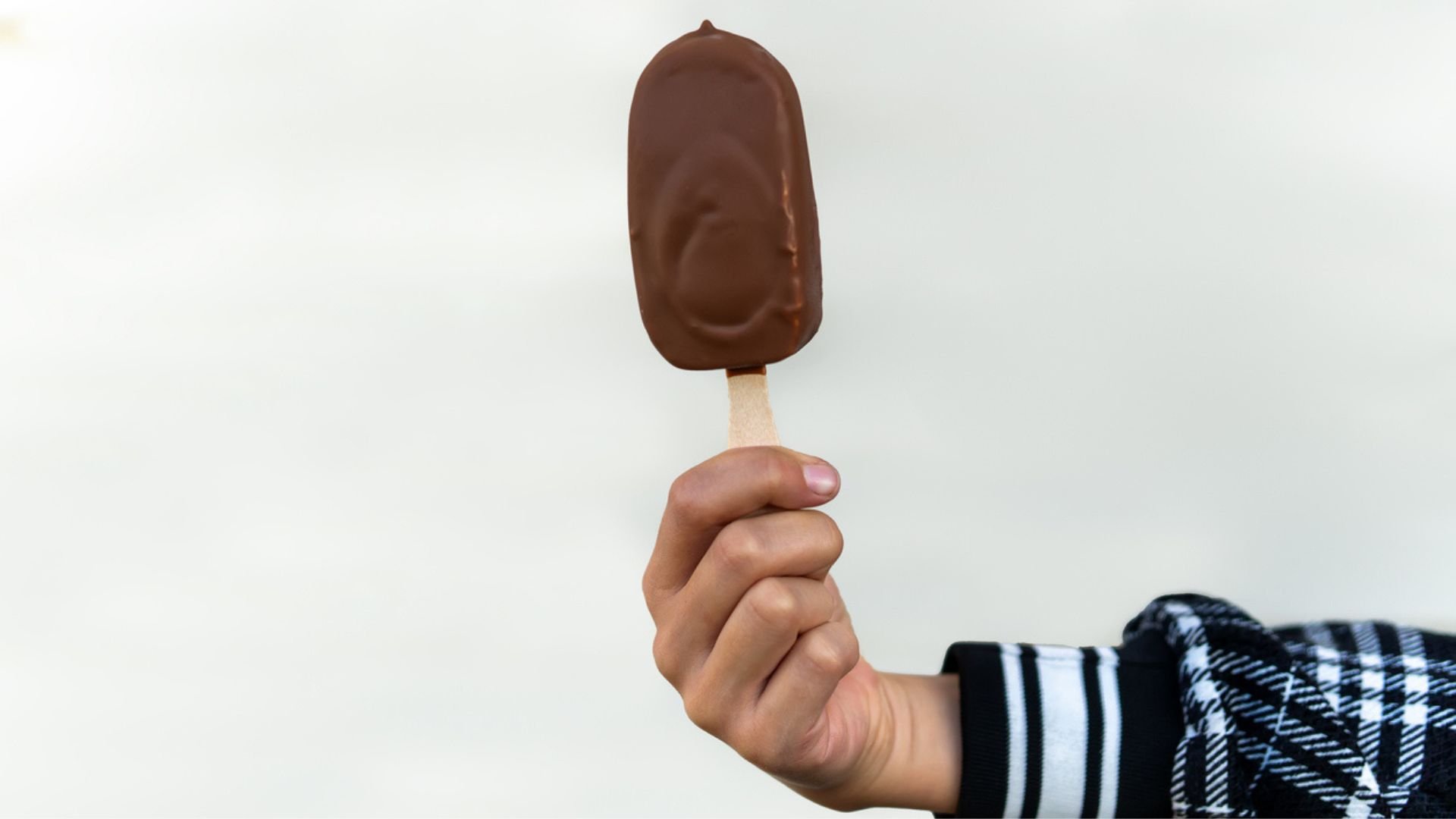 A hand holding a chocolate-covered ice cream bar on a wooden stick against a plain light background. The person is wearing a black and white plaid sleeve.