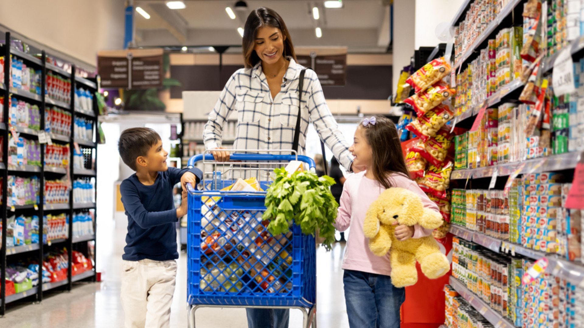 A woman pushes a shopping cart with groceries while smiling at two children, a boy and a girl. The girl holds a yellow stuffed animal as they walk together in a brightly lit grocery store aisle.
