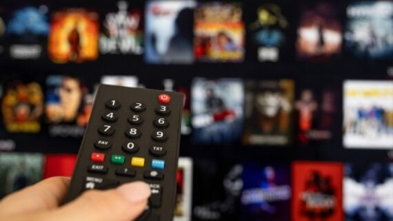 A hand holding a TV remote points at a blurry screen displaying various movie or TV show thumbnails, suggesting the person is selecting something to watch on a streaming service.