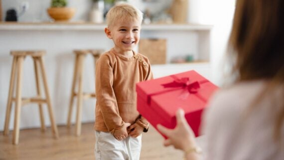 A young boy with blonde hair and a brown long-sleeve shirt smiles excitedly while standing indoors as a person in the foreground holds out a red gift box with a ribbon toward him.