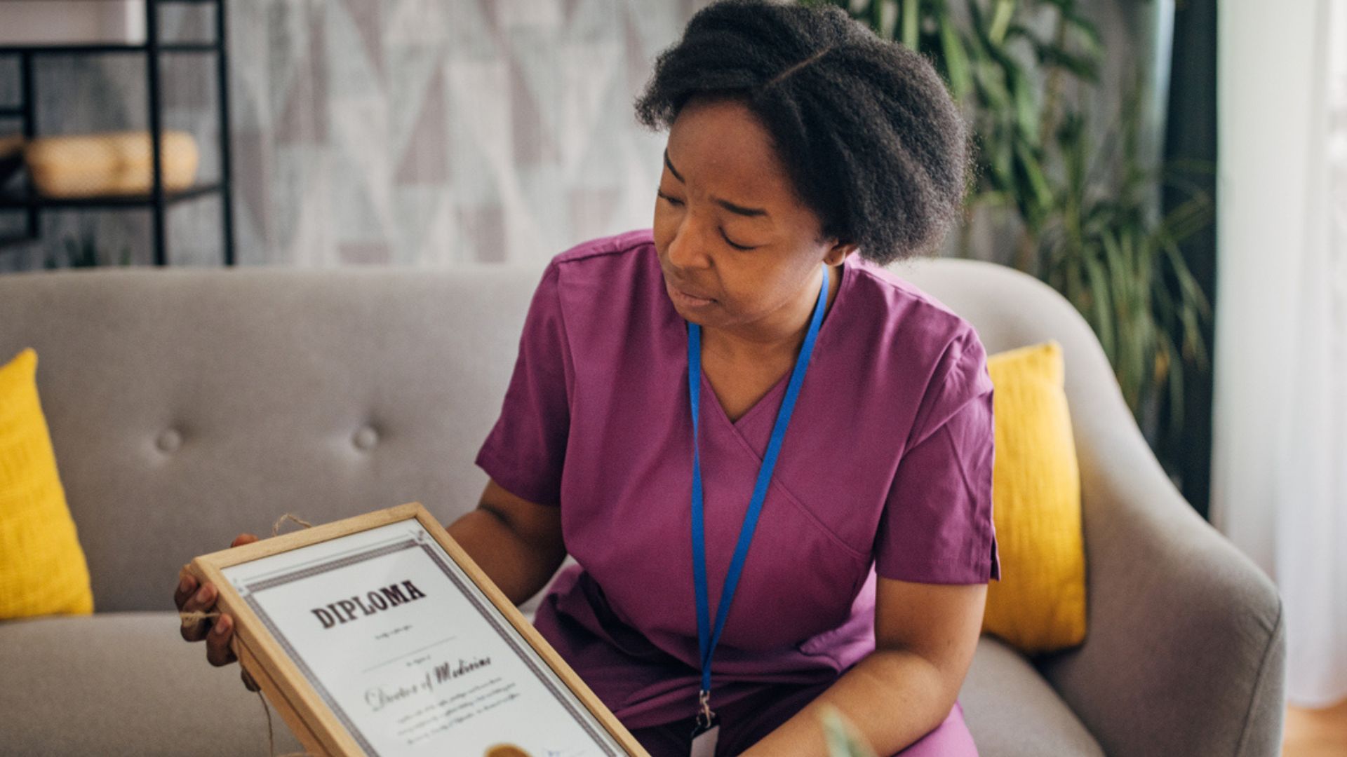 A woman in purple scrubs sits on a sofa and looks thoughtfully at a framed diploma she is holding in her hands. There are yellow pillows beside her and plants in the background.