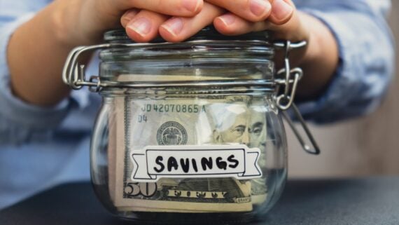 A person places their hands on top of a glass jar filled with U.S. dollar bills, labeled "SAVINGS" with a paper tag. The jar is sitting on a dark surface.