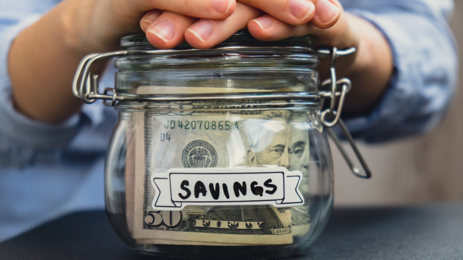A person places their hands on top of a glass jar filled with U.S. dollar bills, labeled "SAVINGS" with a paper tag. The jar is sitting on a dark surface.