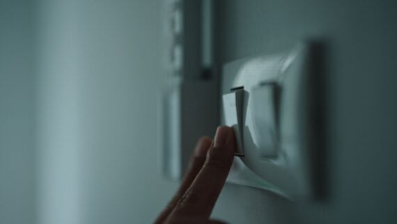 A close-up of a hand reaching to press a light switch on a wall, with a soft, out-of-focus background.