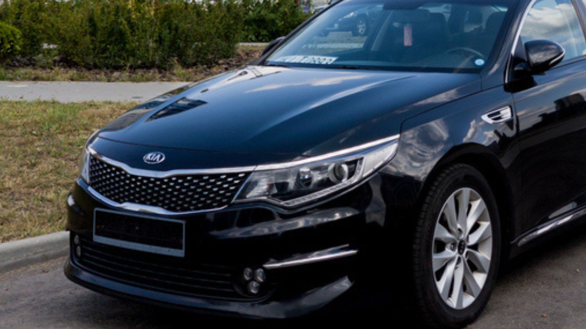 A black Kia sedan is parked on a street next to a curb with grass and bushes in the background. The car is clean and shiny, facing slightly to the right.