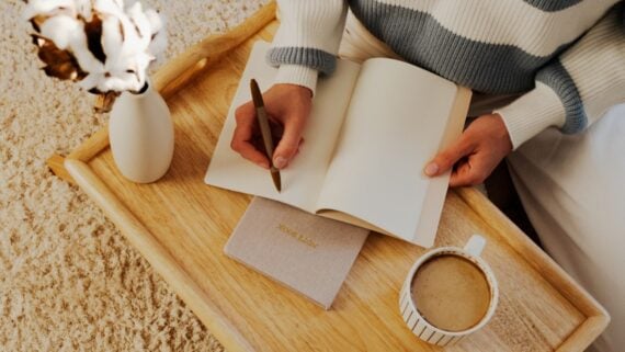 A person sits on the floor with a wooden tray holding an open notebook, a closed journal, a cup of coffee, and a vase with a cotton stem, ready to write while relaxing in a cozy setting.