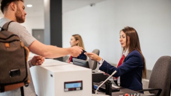A male traveler hands a document to a female airport check-in agent behind a counter, while another agent works at a nearby station in the background.