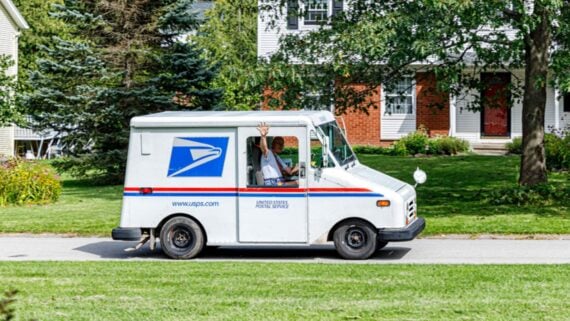 A USPS mail carrier waves from inside a white postal delivery truck as it drives down a suburban street, with green lawns, trees, and houses in the background.