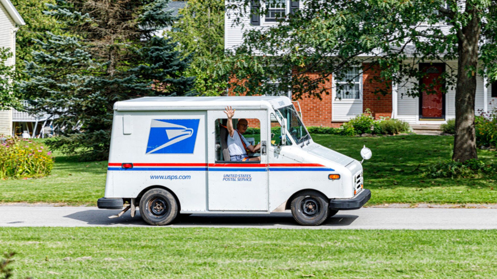 A USPS mail carrier waves from inside a white postal truck while driving through a suburban neighborhood with green lawns and houses in the background.