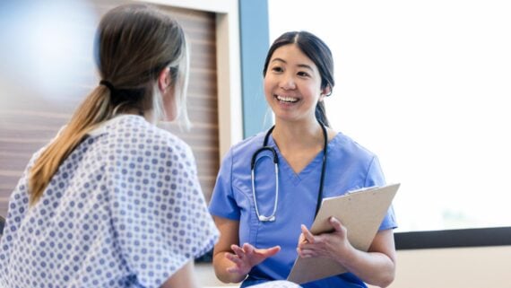 A healthcare professional in blue scrubs with a stethoscope smiles and speaks to a patient in a hospital gown, holding a clipboard during a consultation in a medical office.