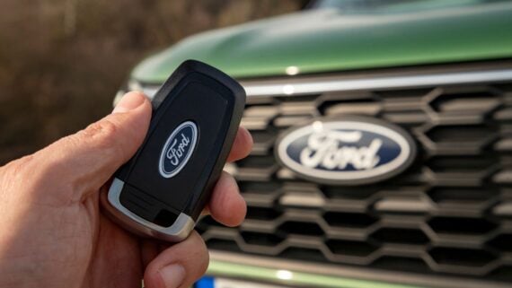 A hand holds a black Ford car key fob in front of the grille and logo of a green Ford vehicle.