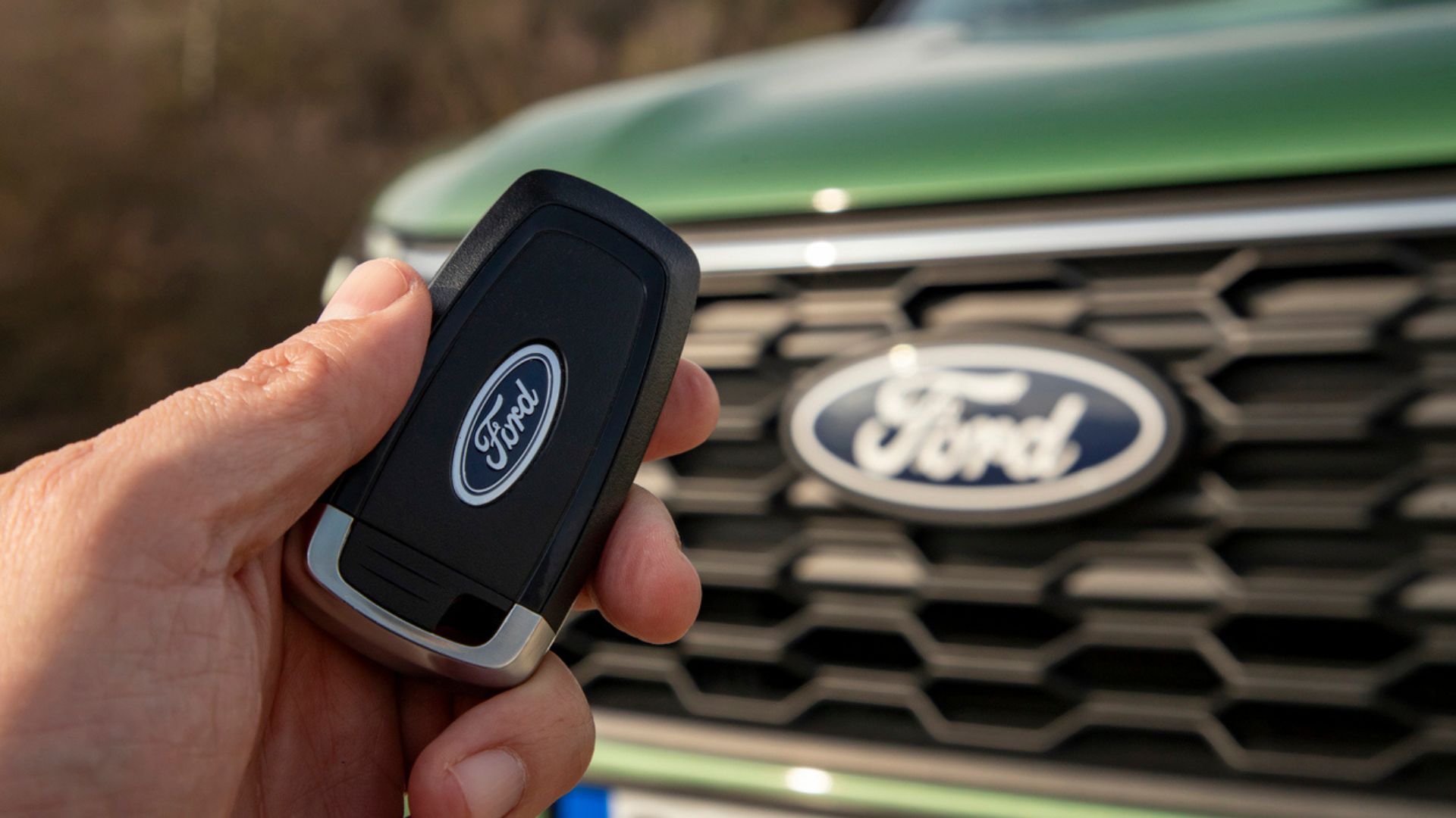 A hand holds a black Ford car key fob in front of the grille and logo of a green Ford vehicle.