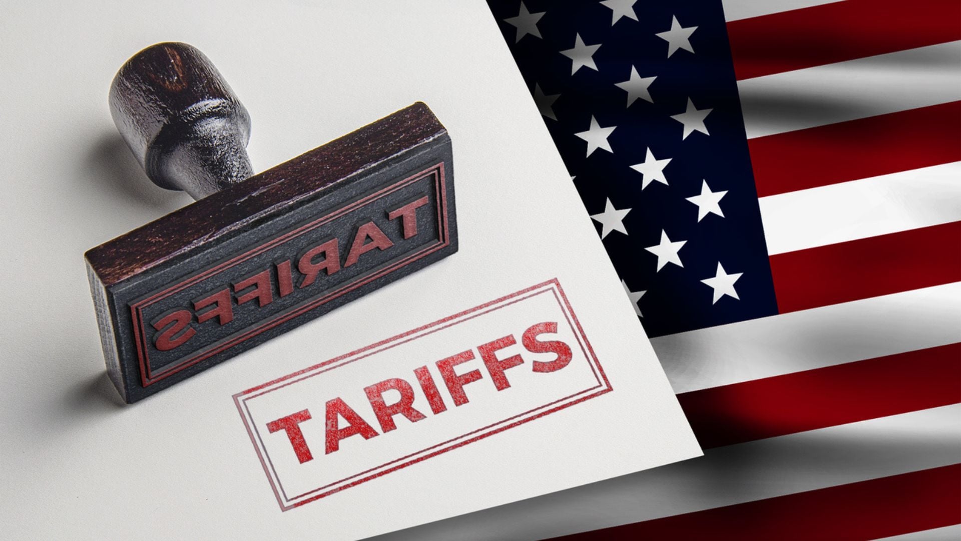 A rubber stamp marked "TARIFFS" lies next to its red imprint on white paper, with the United States flag in the background.