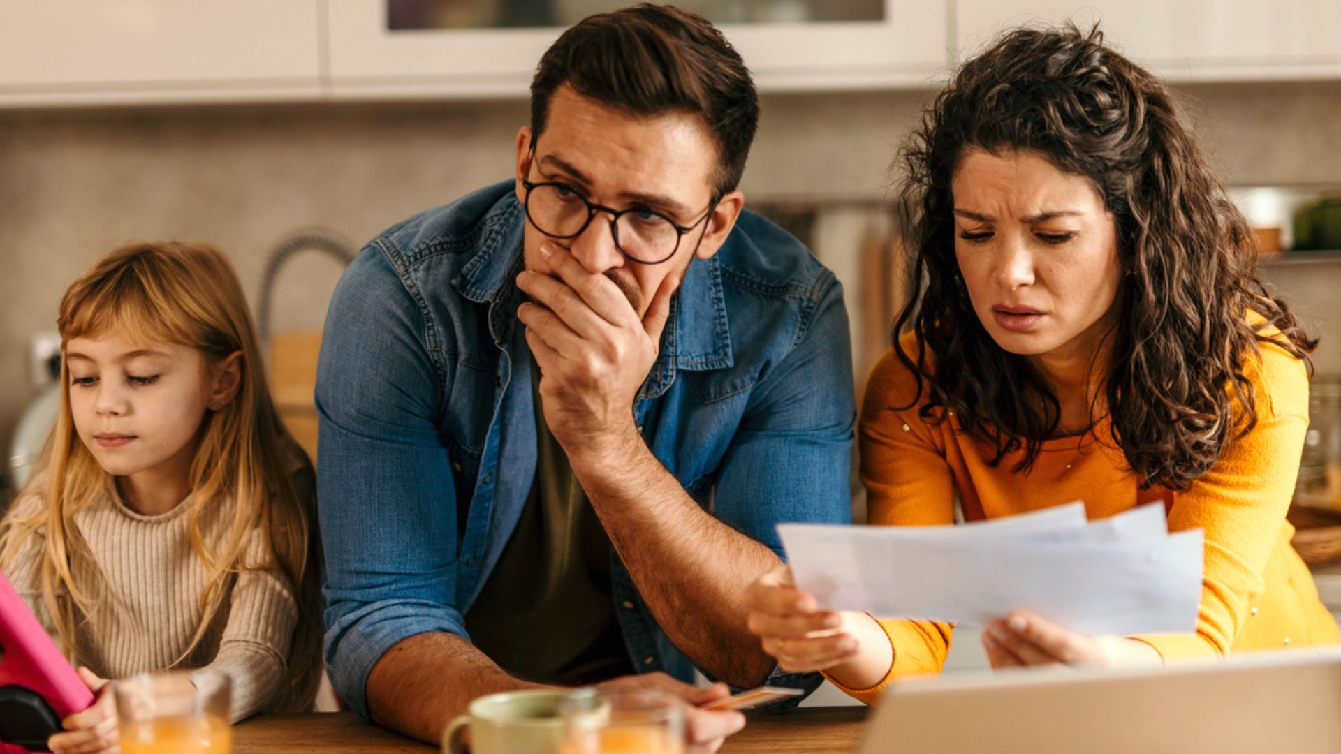 A man and woman look worried while reviewing papers at a kitchen table, with a young girl beside them using a tablet. The scene suggests financial stress or concern over bills.