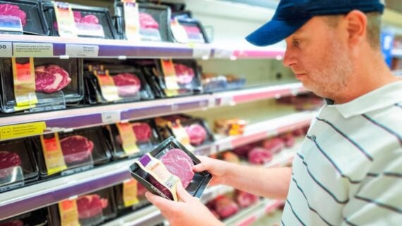 A man in a striped polo shirt and blue cap examines a packaged cut of raw meat in a grocery store meat section, with shelves of various steak packages in the background.