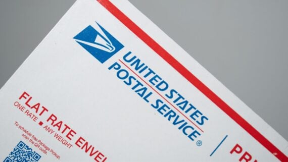 A close-up of a United States Postal Service (USPS) flat rate envelope, showing the USPS logo, blue text, and part of a red border on a gray background.