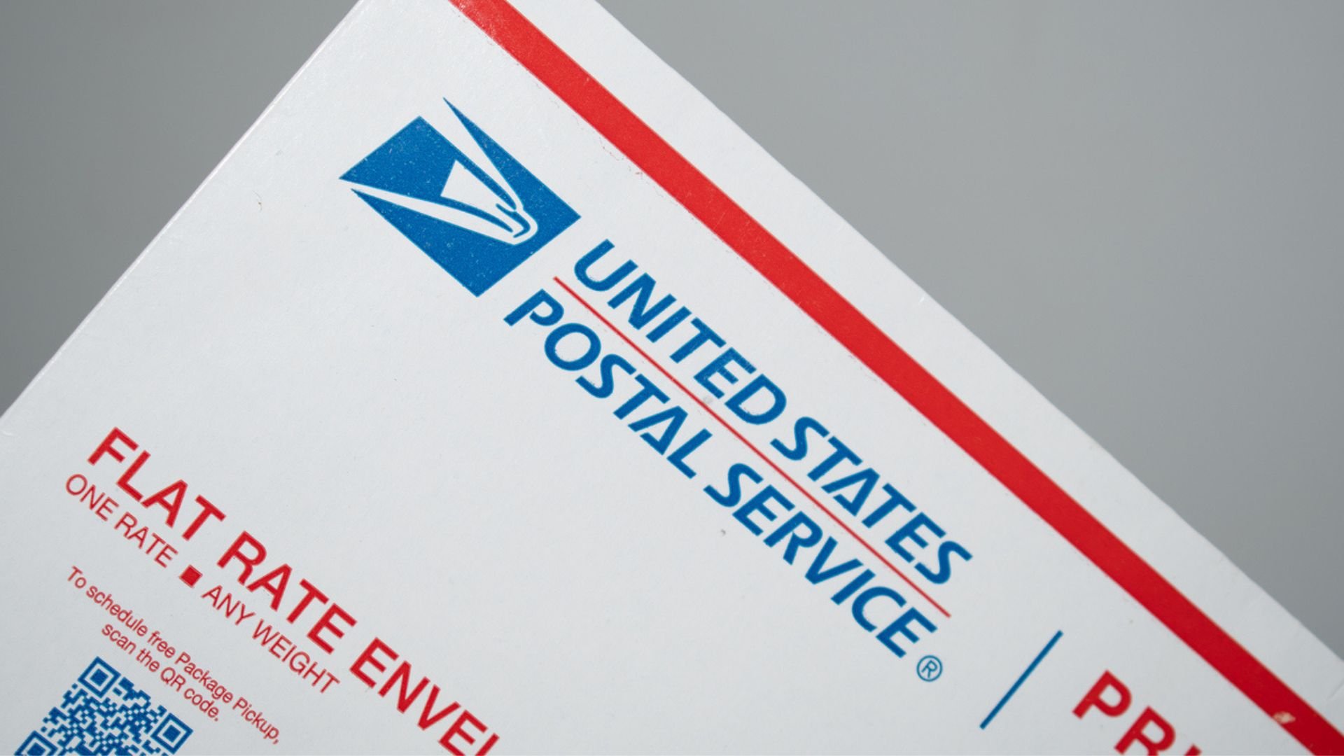 A close-up of a United States Postal Service (USPS) flat rate envelope, showing the USPS logo, blue text, and part of a red border on a gray background.