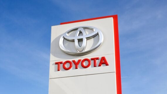 A tall white sign with a red border shows the silver Toyota logo above the word "TOYOTA" in bold red letters, set against a clear blue sky.
