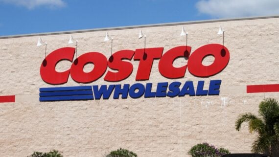 The exterior of a Costco Wholesale store, showing the large red and blue logo sign on a beige brick wall under a blue sky. Shrubs and palm leaves are visible at the bottom.
