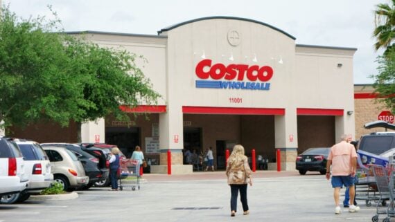 People walk toward the entrance of a Costco Wholesale store, with parked cars and shopping carts visible in the parking lot. The store’s logo is prominently displayed above the entrance.