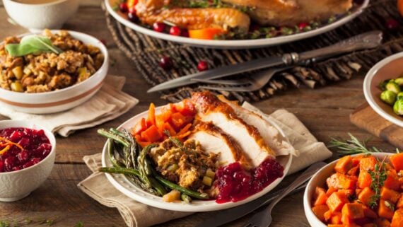 A Thanksgiving meal on a wooden table, featuring sliced turkey, green beans, stuffing, carrots, cranberry sauce, and sweet potatoes, with other dishes and utensils visible in the background.
