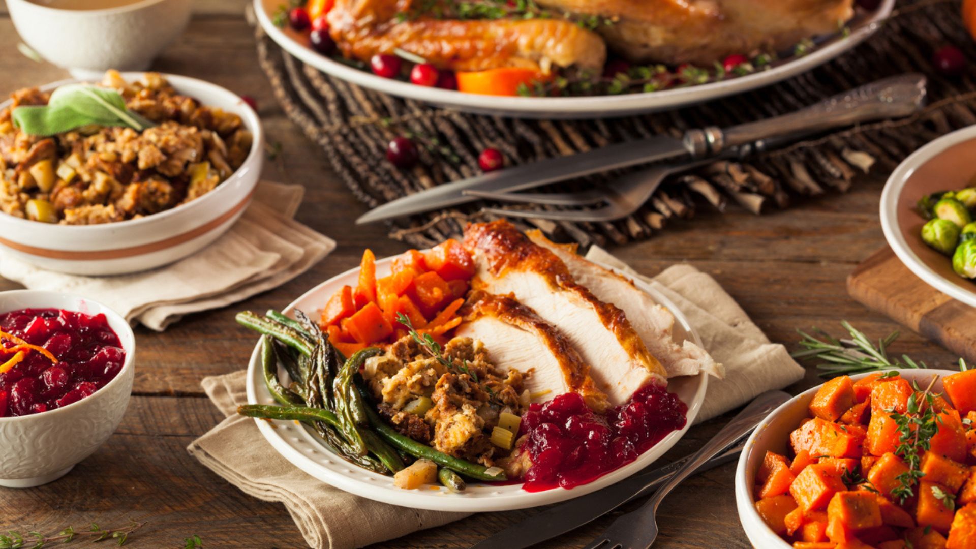 A Thanksgiving meal on a wooden table, featuring sliced turkey, green beans, stuffing, carrots, cranberry sauce, and sweet potatoes, with other dishes and utensils visible in the background.