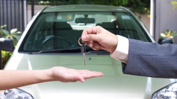 A person in a suit hands over a car key to another person, whose open palm is extended, with a white car in the background.