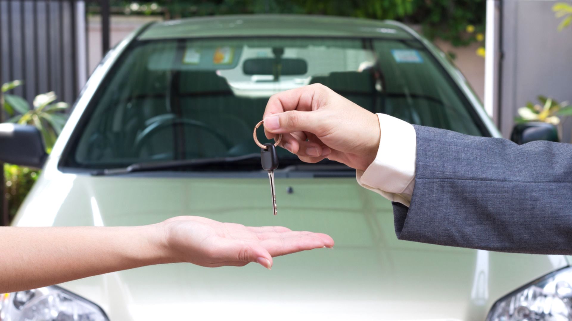 A person in a suit hands over a car key to another person, whose open palm is extended, with a white car in the background.