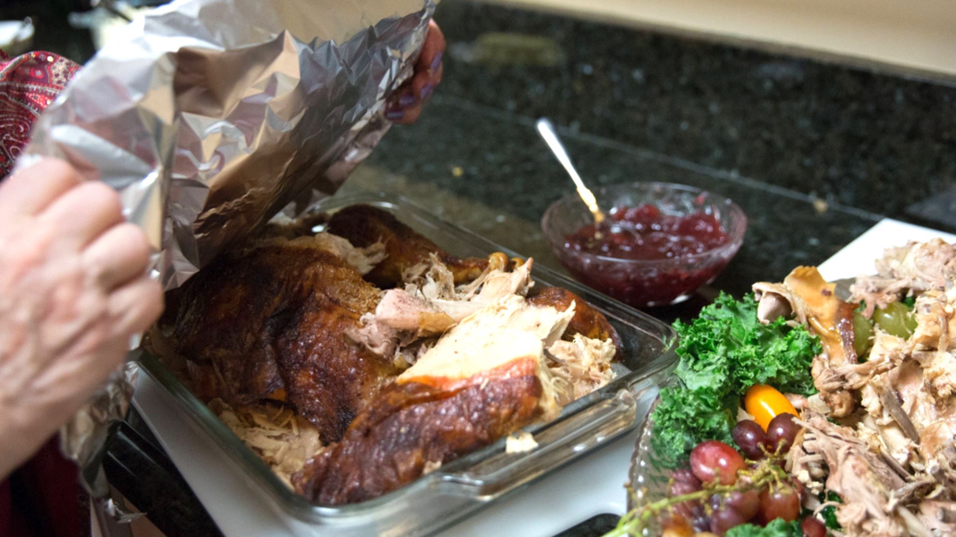 A person lifts foil from a glass dish of roasted turkey on a kitchen counter, with a bowl of cranberry sauce and a platter of turkey, kale, and grapes nearby.