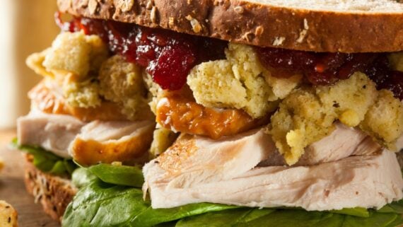 A close-up of a sandwich with sliced turkey, stuffing, cranberry sauce, leafy greens, and oat-topped whole grain bread. The sandwich ingredients are overflowing between the bread slices.