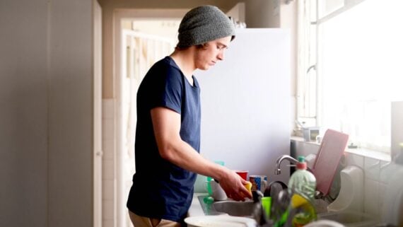 A person wearing a gray beanie and navy t-shirt washes dishes at a kitchen sink, with sunlight coming through the window and various kitchen items on the counter.