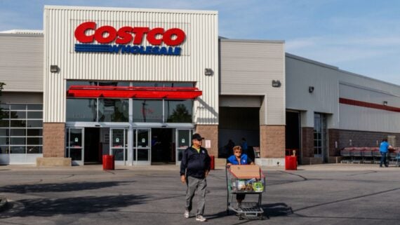 Two people walk out of a Costco Wholesale store with a shopping cart full of groceries on a sunny day. The Costco sign is visible above the entrance.
