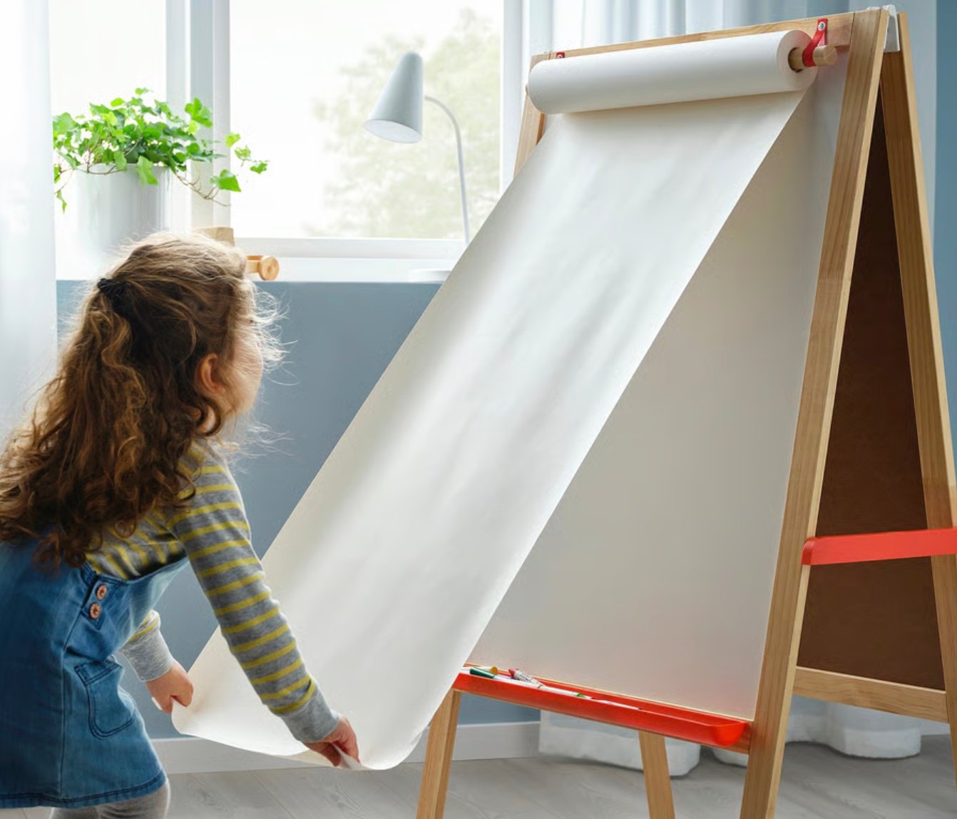 A young child in a striped shirt and denim dress pulls down a large sheet of white paper on a wooden easel in a bright, sunlit room with a plant on the windowsill.